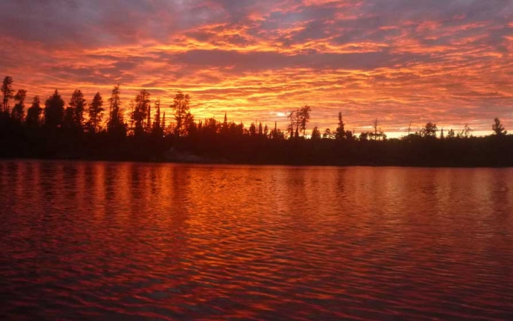 The sky appears in shades of red, orange and purple, reflected in the lake below, between a line of trees. 